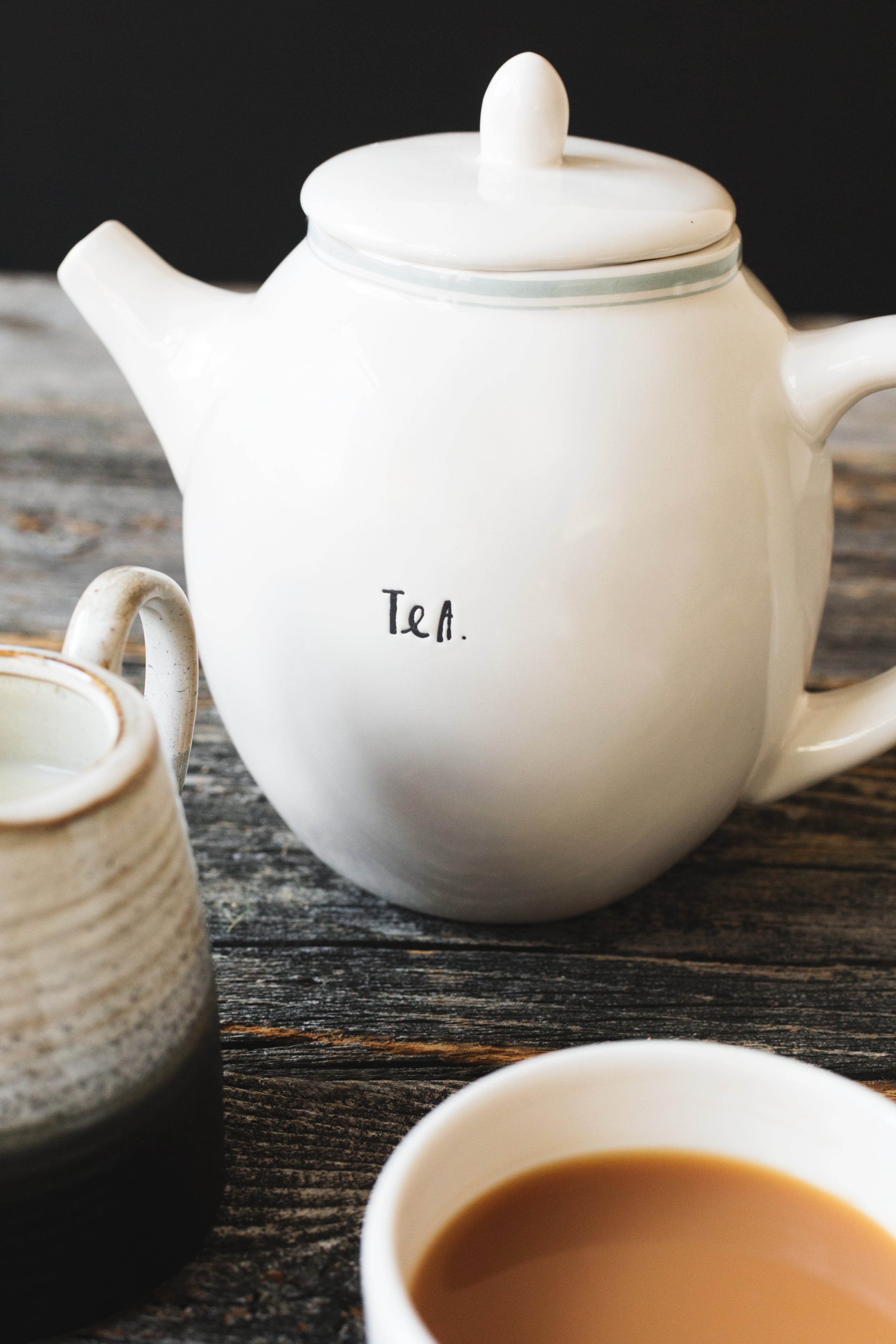 A white teapot hand-lettered “Tea.” beside a cup of milky tea on a weathered wooden table